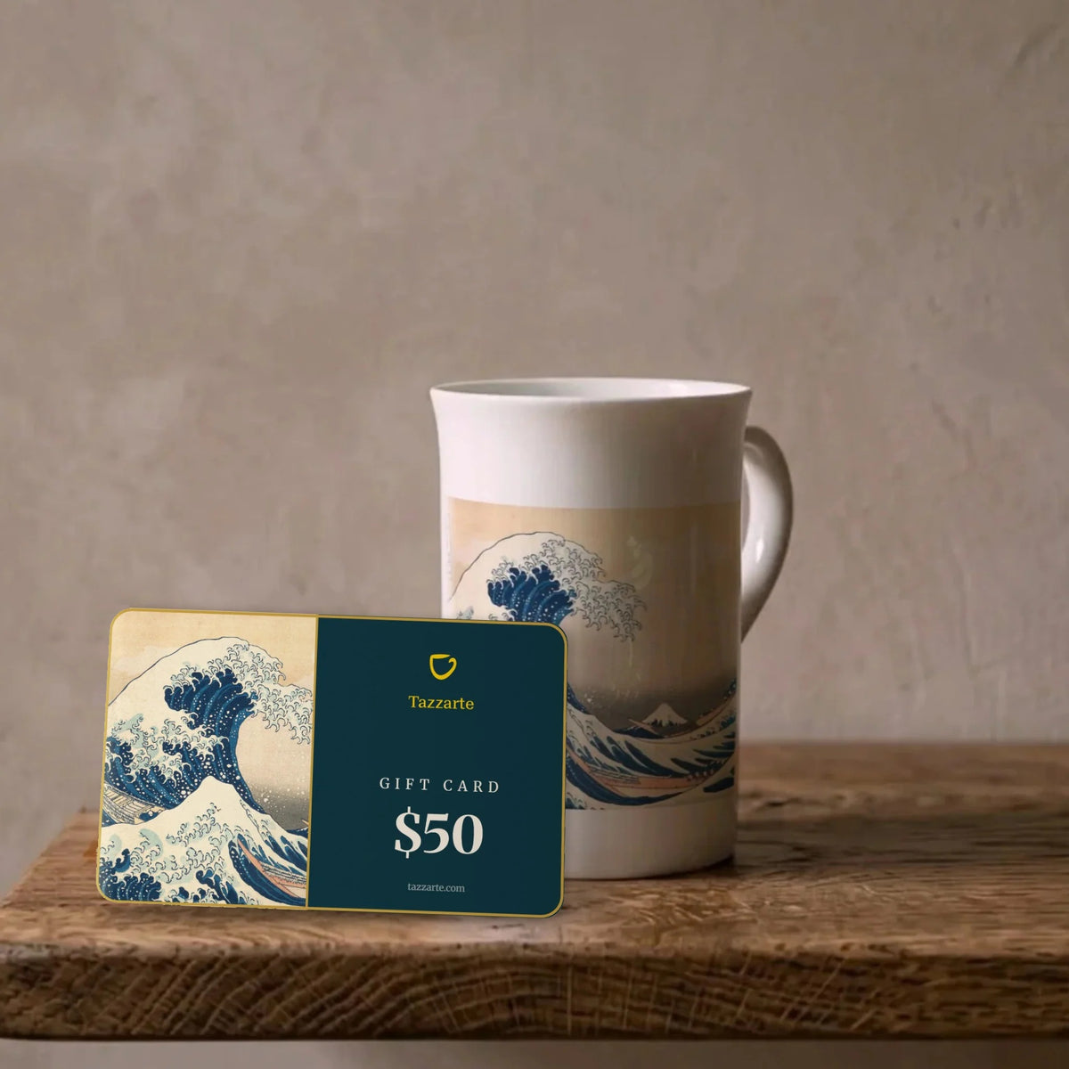 Tazzarte-Kunstgeschenkkarte über $50 mit Hokusais Großer Welle neben der passenden Kunsttasse aus Porzellan auf einem Eichenregal
